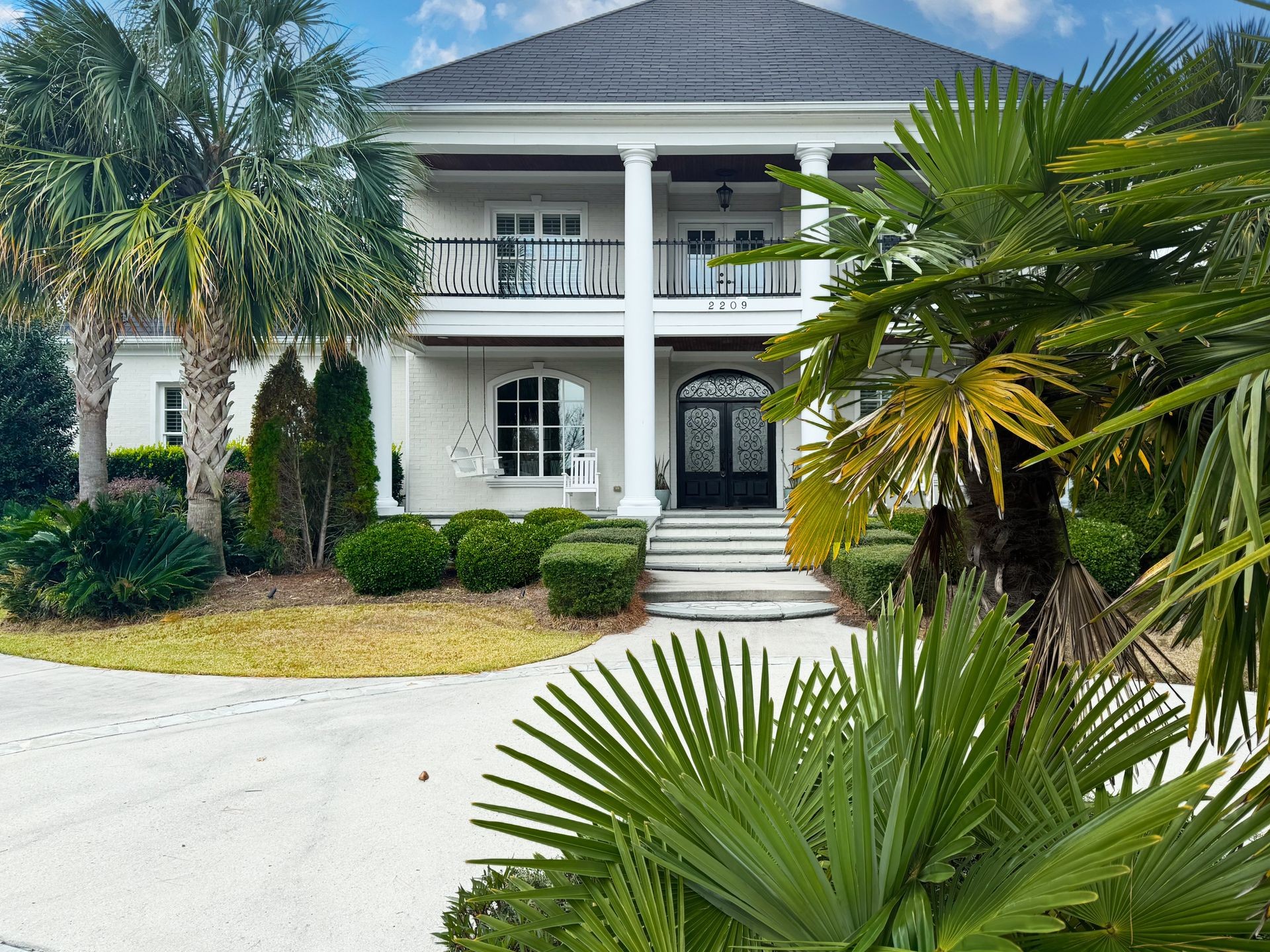 Elegant white two-story house with columns and a balcony, surrounded by lush greenery and palm trees.